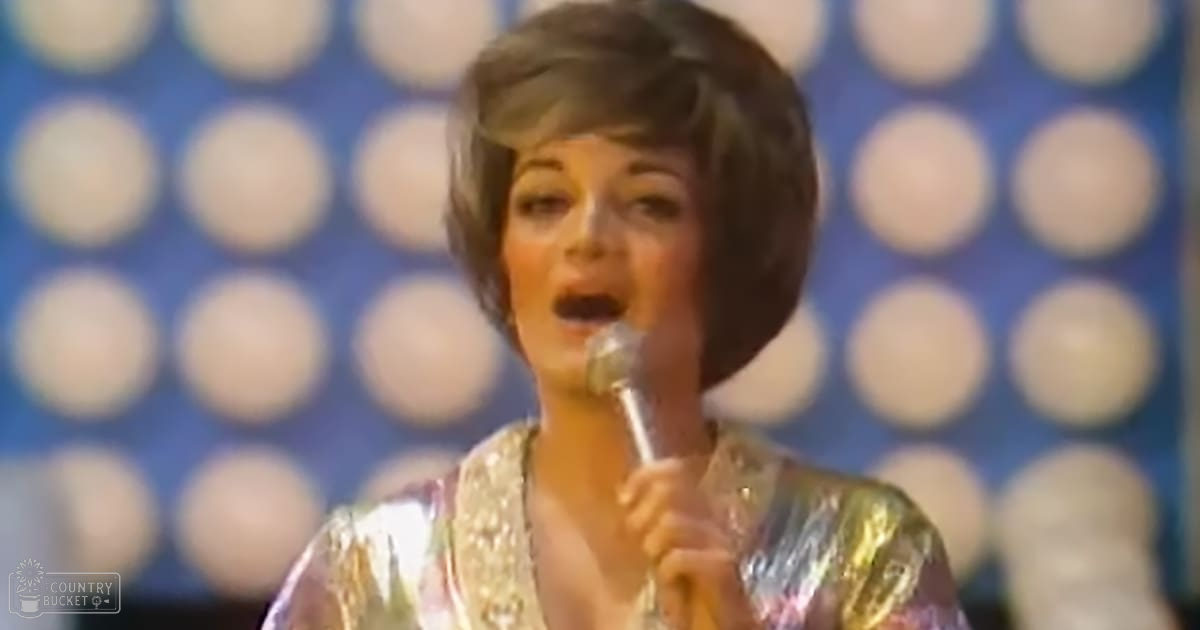 An adult female performer in a sparkly, sequined 1960s-style top sings into a microphone against a blue backdrop with circular patterns, likely in a television or recording studio.