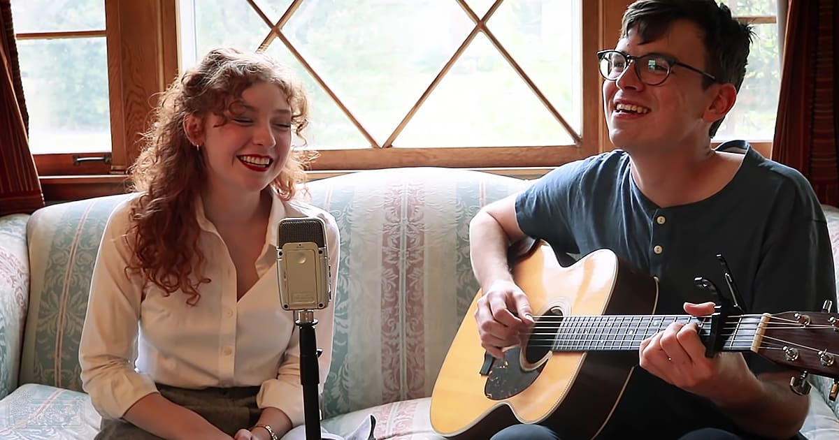 A young adult female with curly hair in a white button-up shirt, alongside a young adult male with short hair wearing glasses and a dark blue henley shirt. They are performing music together indoors, possibly in a living room.