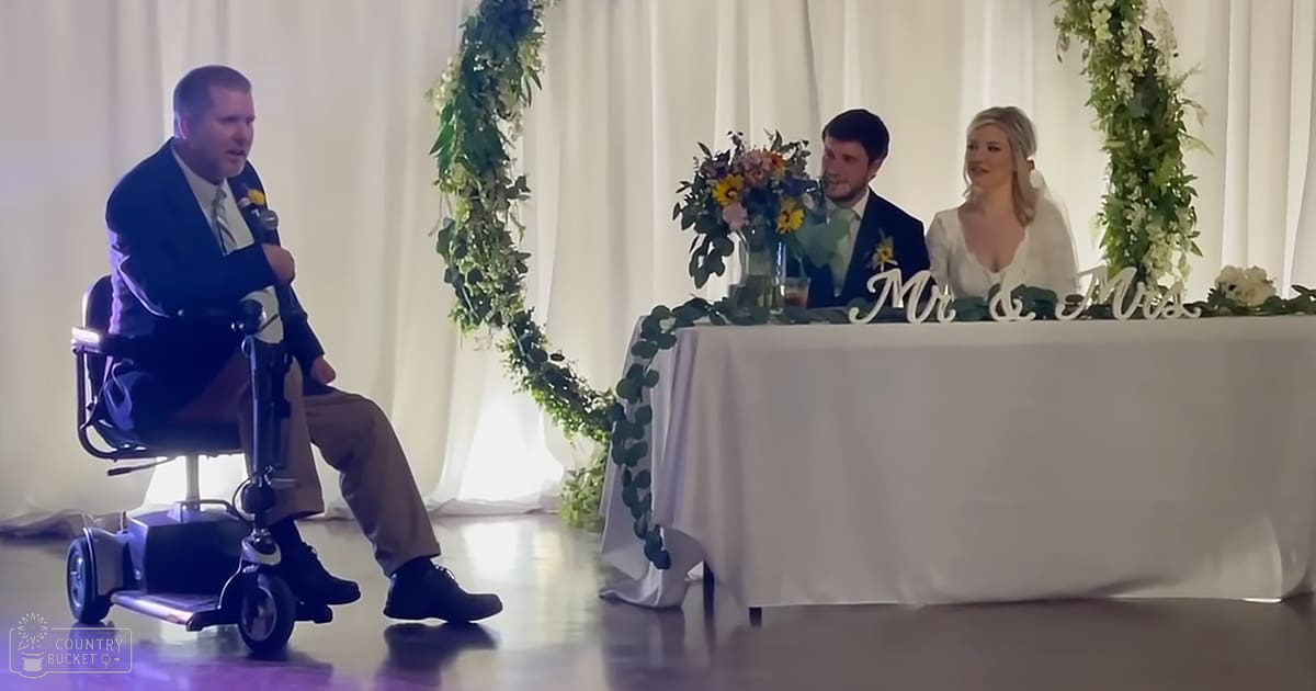 A wedding speech at an indoor event with a couple as the bride and groom seated next to each other. The setting includes white linens, floral arrangements, greenery decorations, and a sign reading 'Mr & Mrs'.