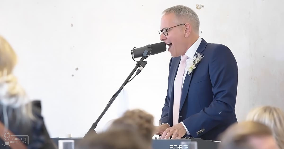 A middle-aged man giving a speech at an event, possibly a wedding reception, with a microphone stand and text 'AD-8' visible in the foreground. The setting is indoors with plain white walls and natural daylight.