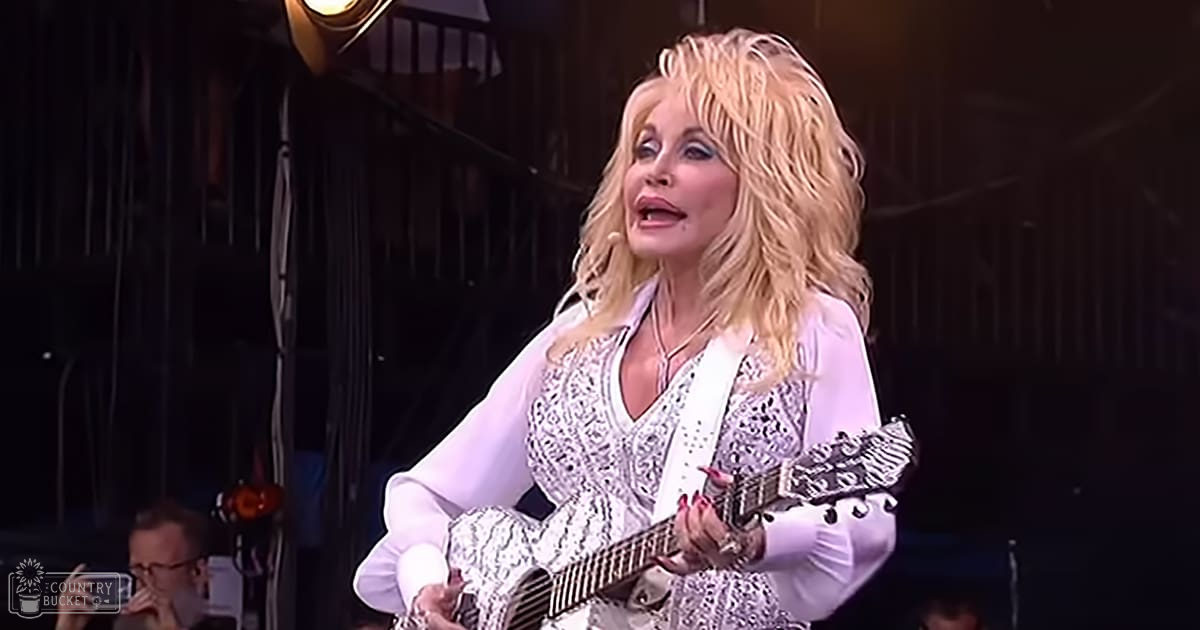 A middle-aged to older adult female performer in a white blouse with intricate lace details, performing at an outdoor concert venue under artificial lighting.