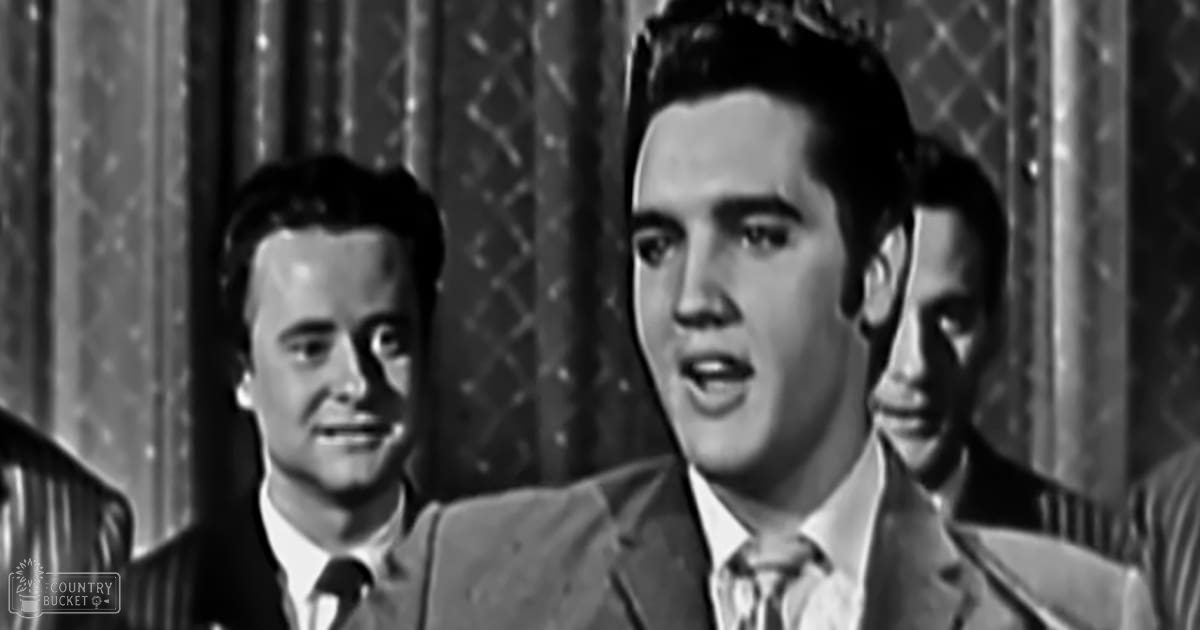 A young adult male wearing a mid-20th-century suit jacket sits in an ornately decorated indoor setting with curtains, surrounded by two other similarly dressed individuals.
