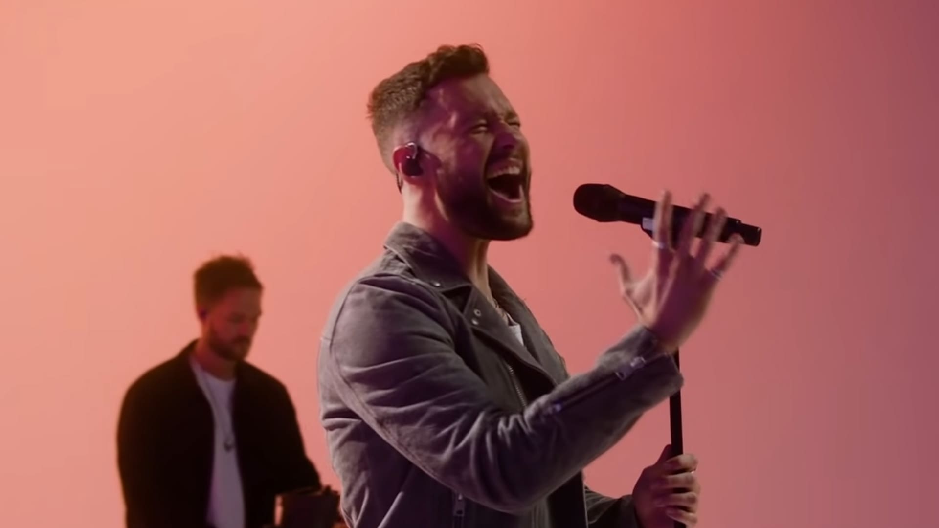Young adult male performing live on stage, wearing a dark gray jacket over light-colored shirt and black pants with microphone stand in foreground; background is solid pink hue from stage lighting.