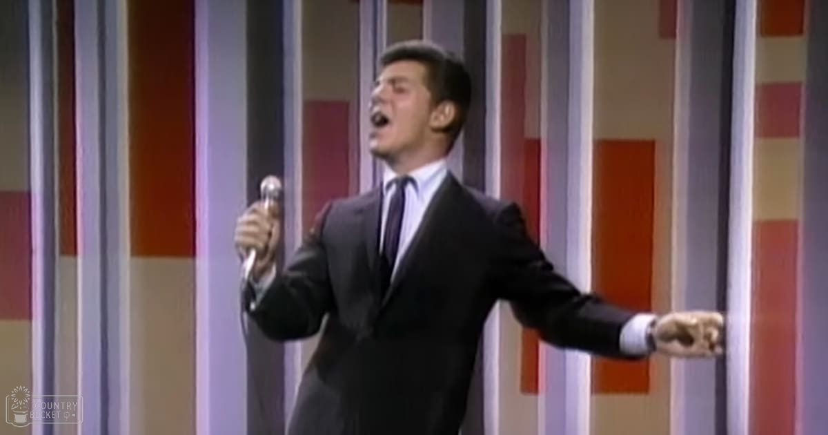 A young adult male singer wearing a dark gray formal suit with white shirt and tie, performing on stage in a studio setting.