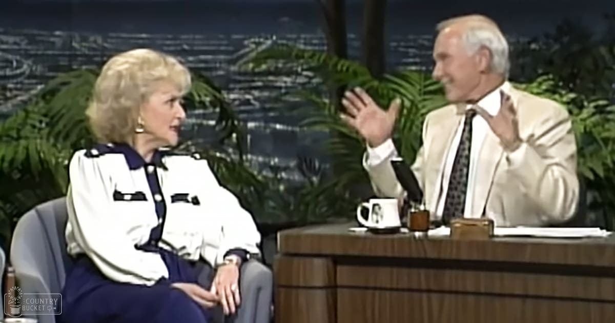 Two adults, a middle-aged to older female wearing a white blouse with dark blue accents and buttons, paired with dark pants or skirt, and an older male in a light-colored suit jacket with a dark tie and white shirt underneath, seated at a wooden desk in a talk show setting with greenery and cityscape background.