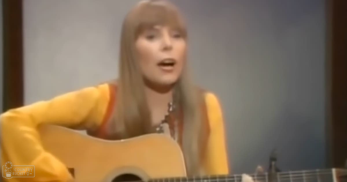 A young adult female plays an acoustic guitar in a studio setting, wearing a yellow garment with wide sleeves and a low-cut neckline. The lighting is even and bright.