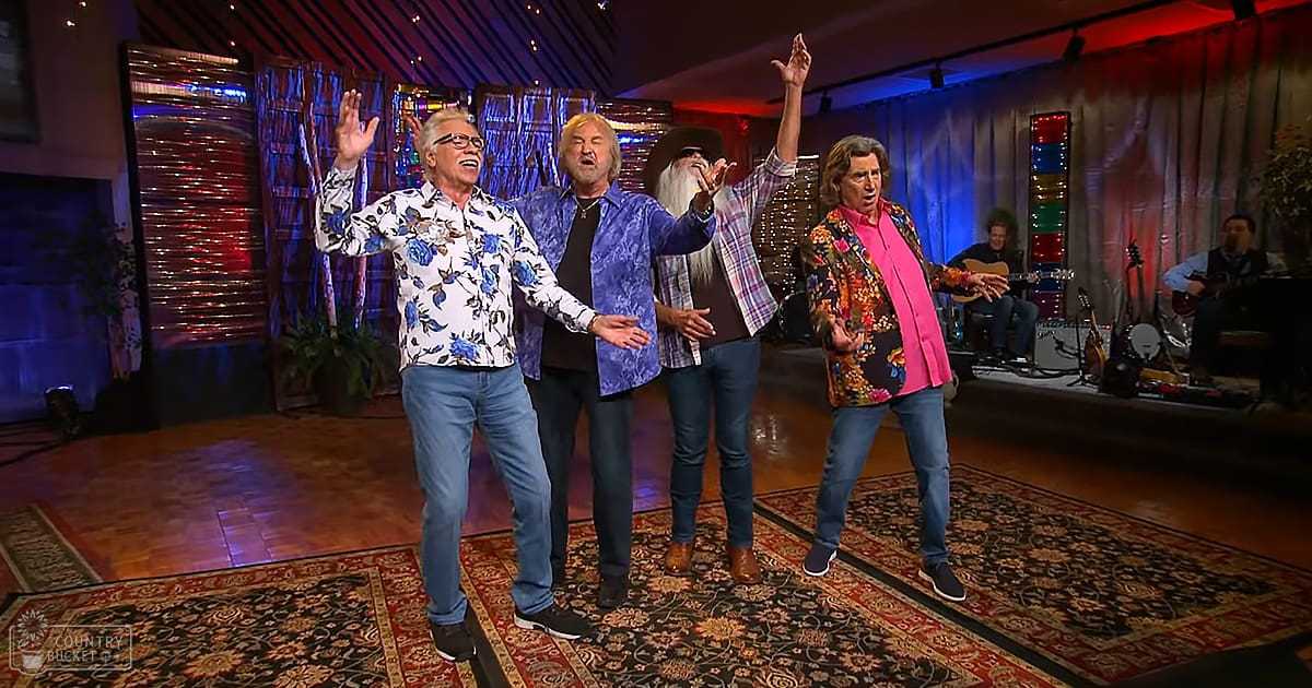 Adult male singer wearing a white shirt with blue floral patterns, performing alongside others in an indoor stage or studio setup with patterned rugs and decorative lighting.