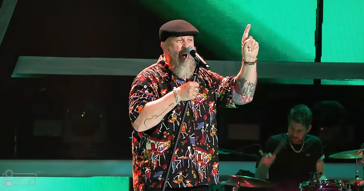 A middle-aged male performer wearing a colorful, instrument-patterned shirt and hat at an indoor concert venue with stage lighting.