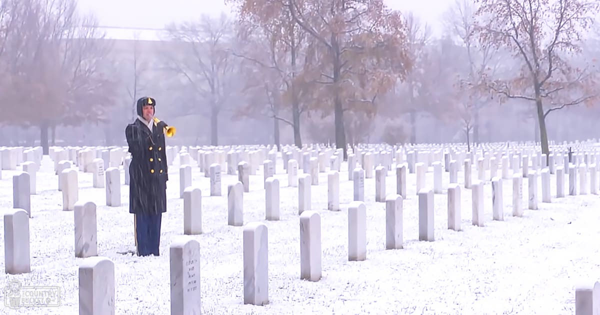 An adult male singer in a winter cemetery playing a yellow bugle, dressed in a dark blue military-style jacket with gold buttons and a matching cap.