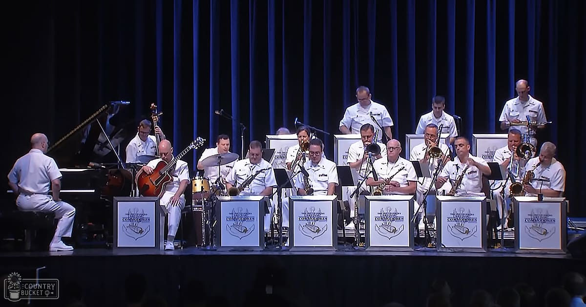 A military band performs on stage at an indoor concert venue with dark blue curtains, sheet music stands displaying 'The Navy Pipers Commissaries' logo.