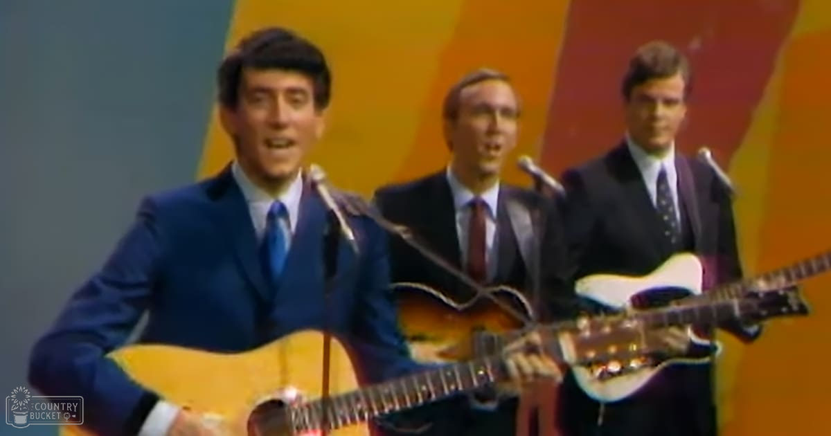 A young adult male singer in a blue suit jacket and white shirt with a blue tie stands on stage, accompanied by two other young adult males wearing similar formal attire.
