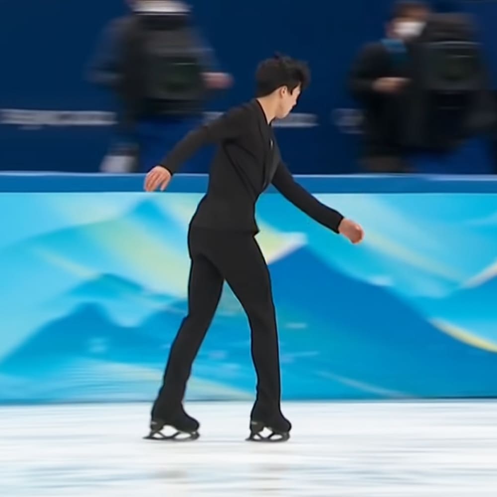 A young adult male figure skater in a black long-sleeve skating outfit, ice skating at an indoor rink with spectators and blue abstract designs on the background.