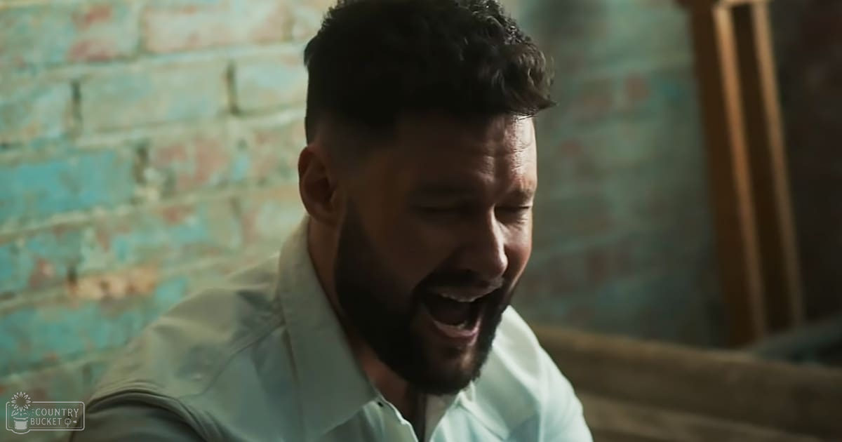 An adult male with short dark hair and a beard wearing a light blue collared shirt, set against a rustic indoor backdrop with painted brick walls, suggesting daytime natural lighting.