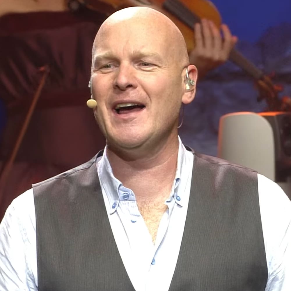 A middle-aged male in a relaxed fit white shirt layered under a dark gray vest stands against an indoor studio backdrop with musical instruments and a microphone headset, suggesting he might be performing or speaking.