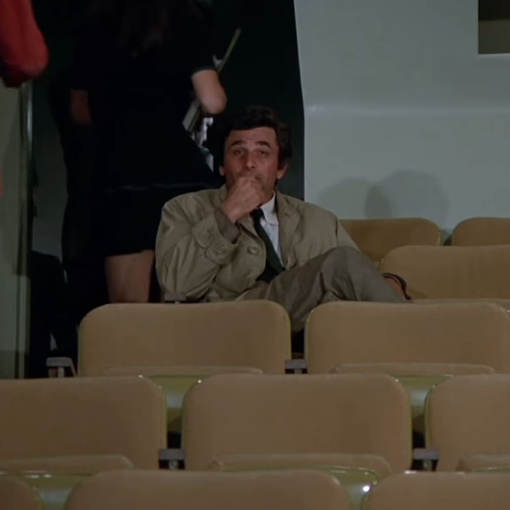 A middle-aged man in a beige trench coat stands barefoot or wearing something not visible, surrounded by rows of beige chairs in an indoor auditorium setting with artificial lighting.