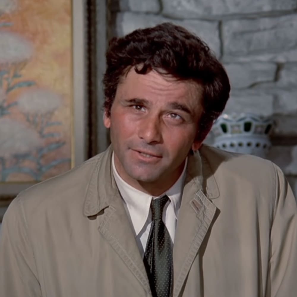 A middle-aged adult male wearing a light beige trench coat with double-breasted front and lapels, standing indoors against a brick wall backdrop with natural daylight.