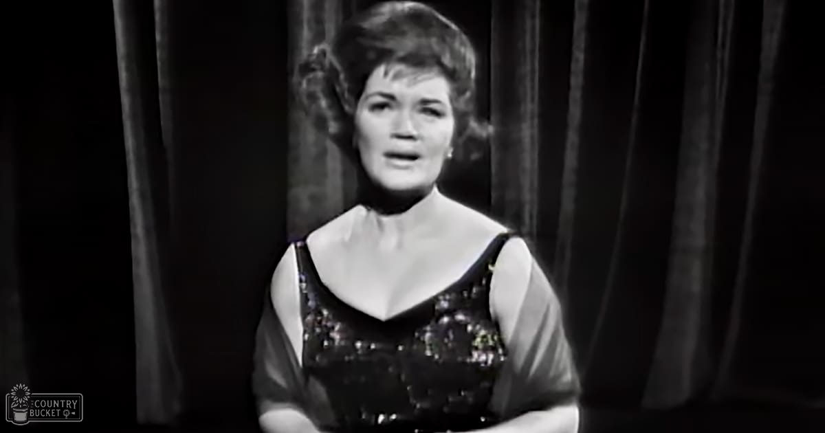 Adult female wearing a sleeveless, fitted dress with high neckline and short sleeves, standing against dark curtains suggesting an indoor stage or performance area.