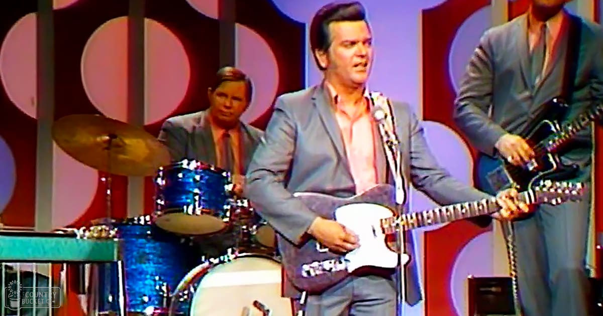 A mid-20th century male performer wearing a light gray suit jacket and white shirt, accompanied by other musicians in formal attire, set against a vibrant stage backdrop with large circular patterns.