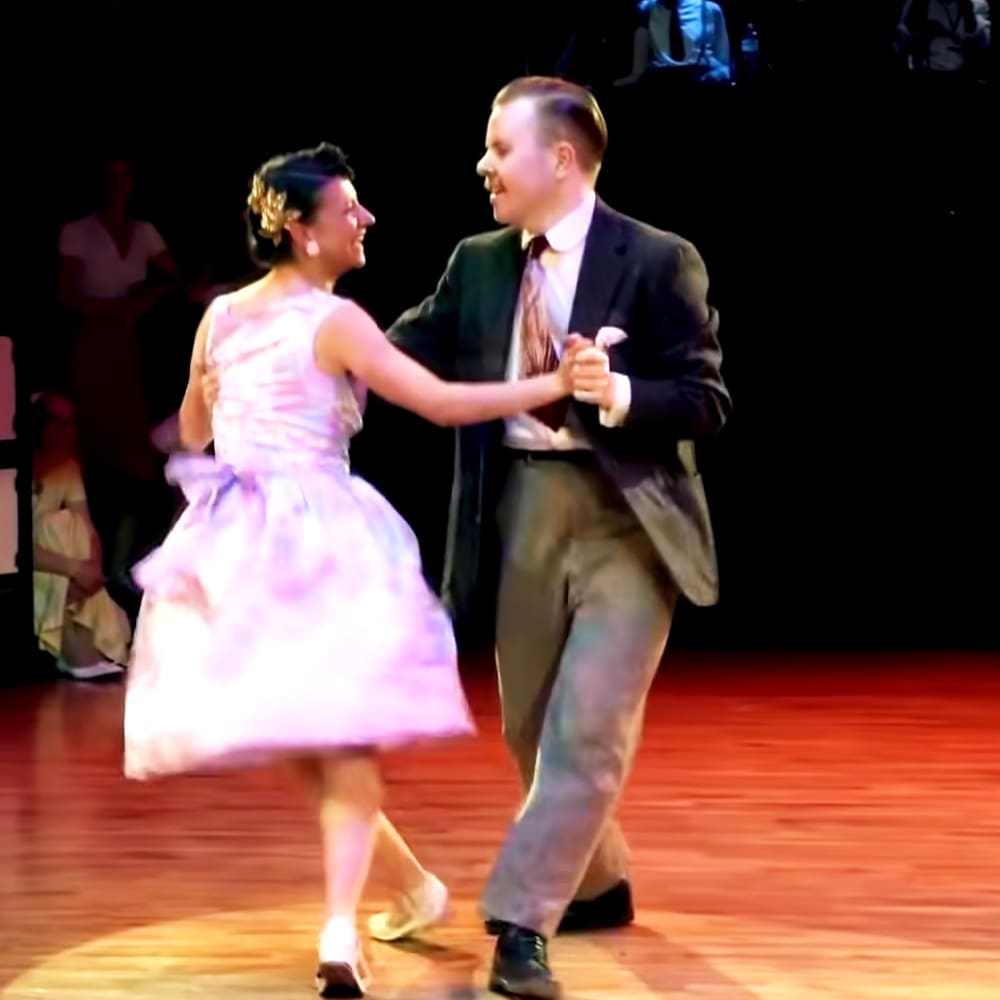 A couple dancing in a ballroom setting, with the woman wearing a light pink floral dress and white heels, and the man in a dark suit jacket and black dress shoes.