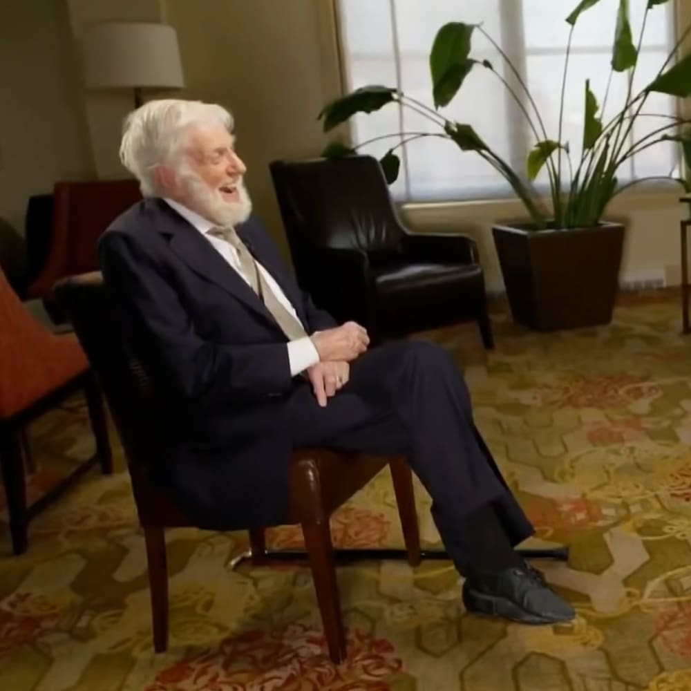 An older adult male in a well-furnished hotel room with natural light, wearing a dark blue suit jacket, white dress shirt, and light-colored tie, surrounded by furniture like an armchair and a floor lamp.