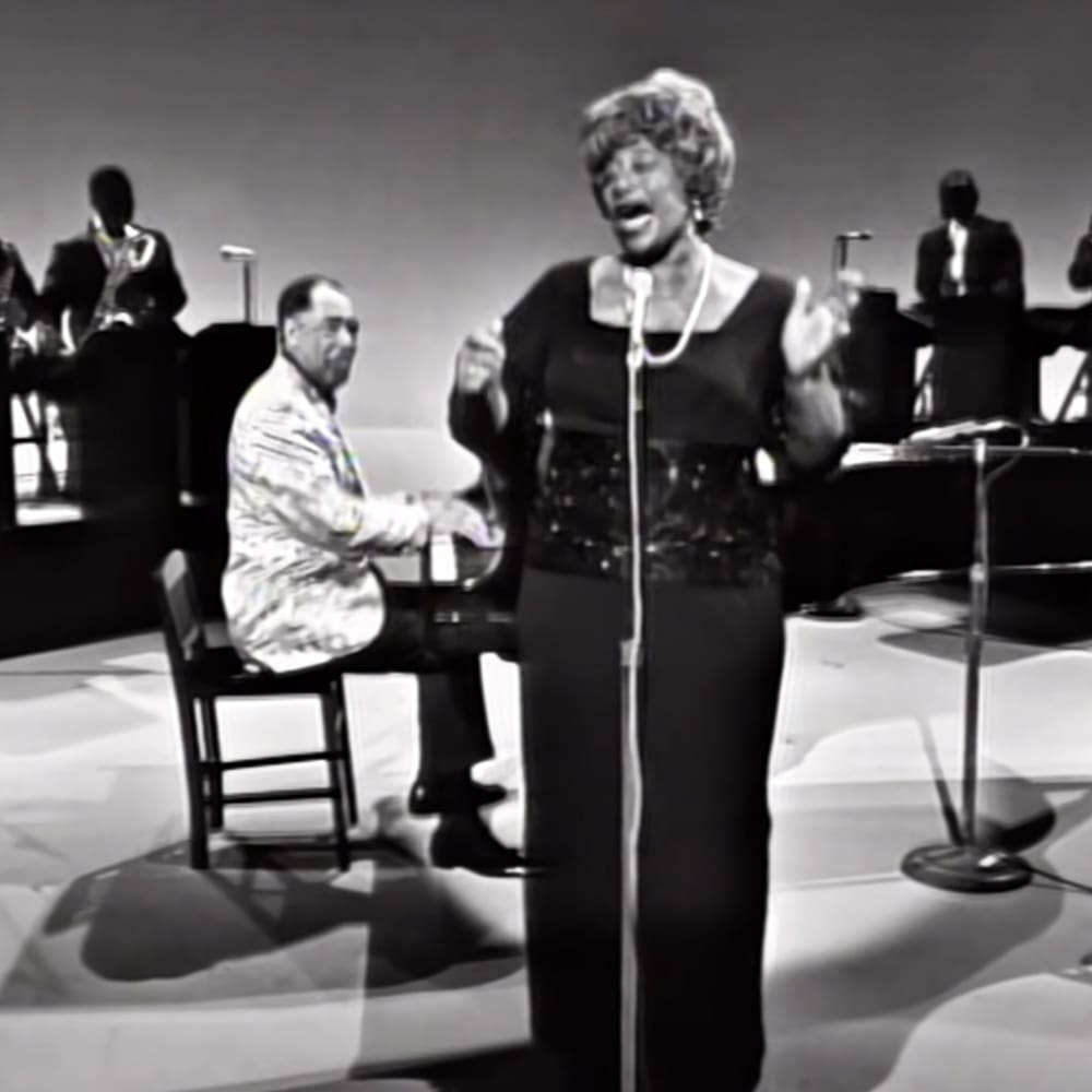 A woman singing on stage, wearing a textured black dress with long sleeves, accompanied by an older adult male pianist in a patterned light-colored suit jacket and dark trousers.
