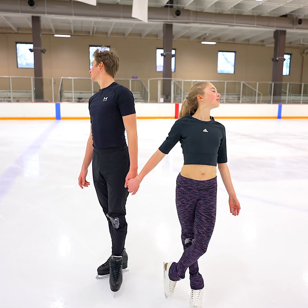 A young couple skating on an indoor ice rink, wearing athletic attire.