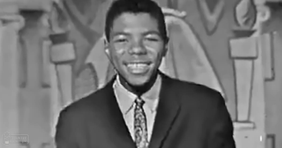 A young adult male wearing a dark suit jacket with a patterned tie stands against an ornate backdrop suggesting a formal indoor setting, likely from the mid-20th century.