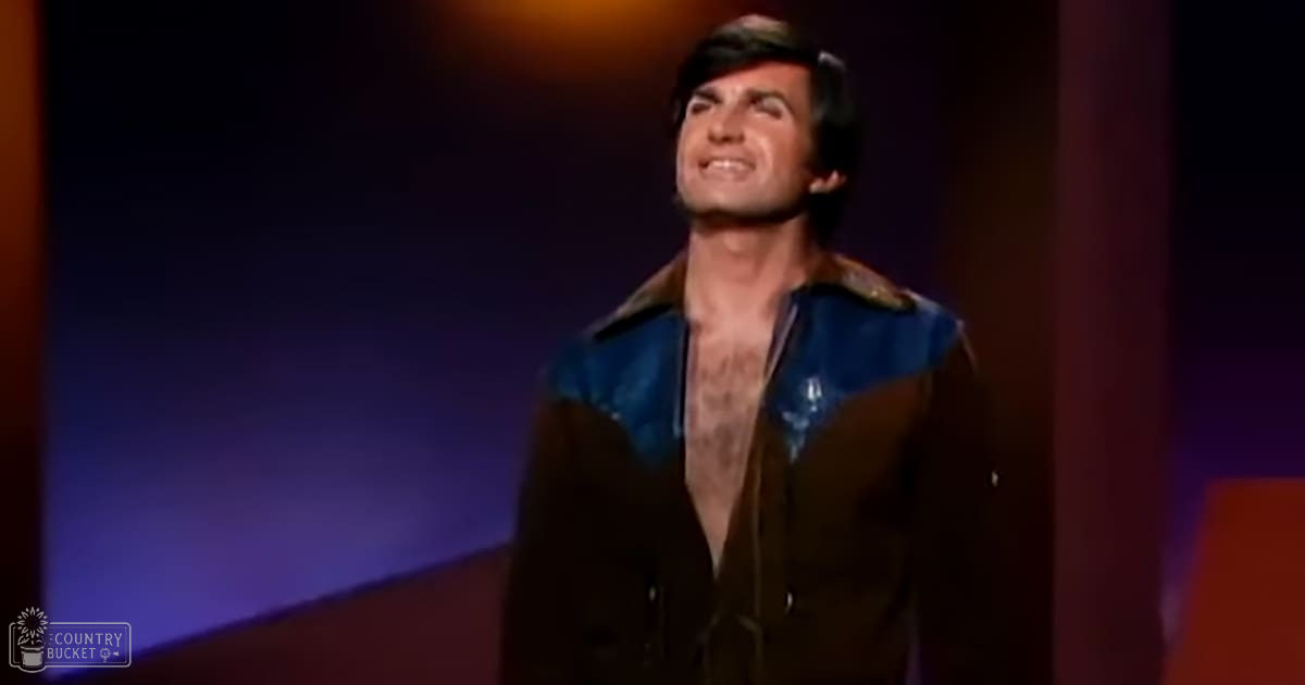 Adult male performer in a loose-fitting brown jacket with blue accents, standing on an indoor stage under purple-orange gradient lighting.