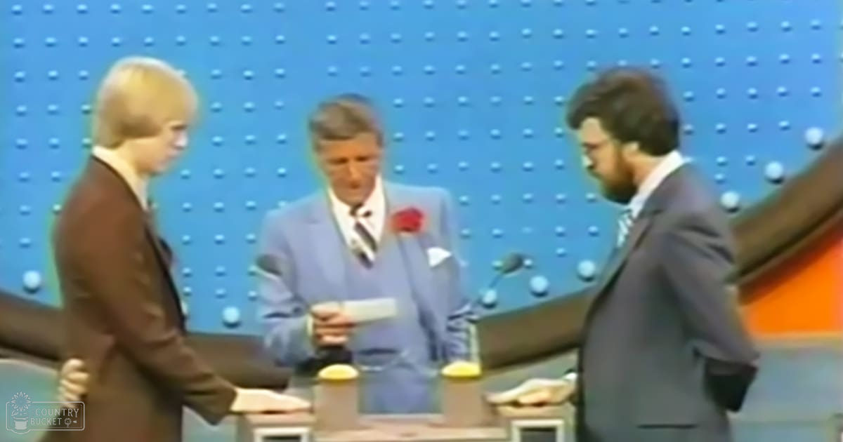 A game show set with three adult male hosts in formal mid-20th century attire, seated at a table with microphones and a small yellow object on it against a blue pegboard wall backdrop.