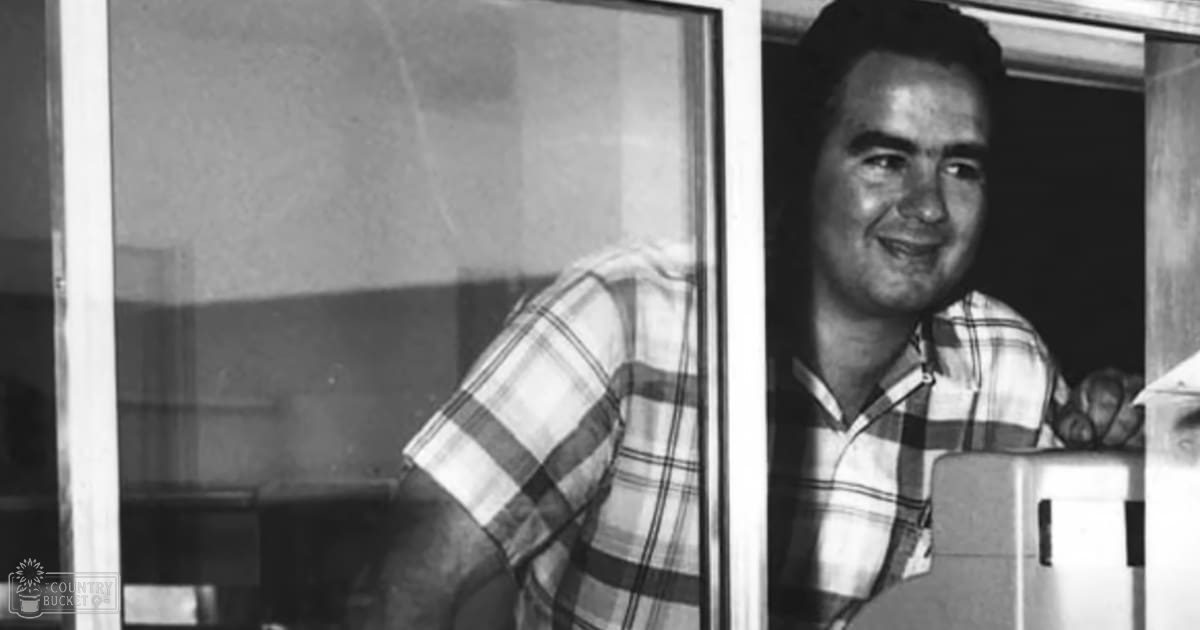 A middle-aged adult male wearing a relaxed fit plaid short-sleeved shirt with white and dark stripes, indoors in what appears to be an office or study area, natural light through window, daytime scene.