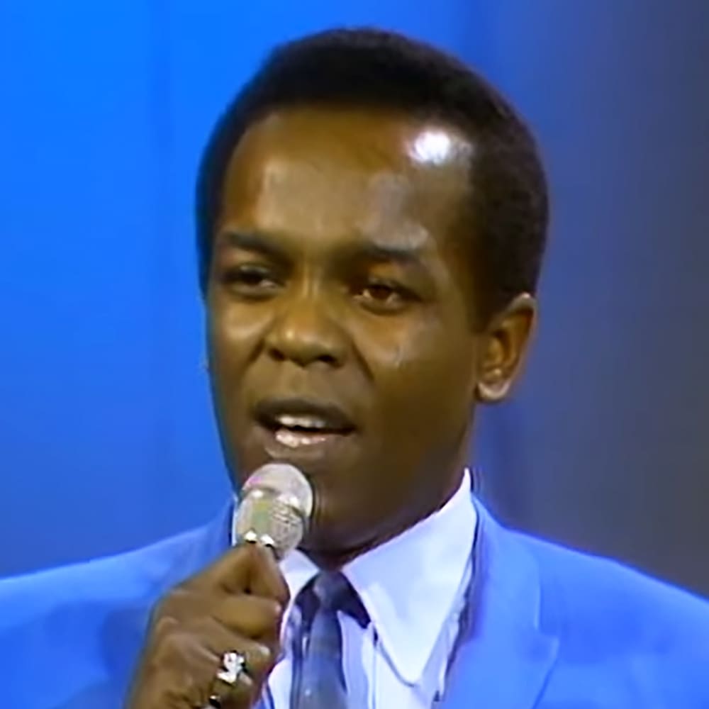 A young adult male in a tailored blue suit jacket with a white shirt, holding a microphone indoors.