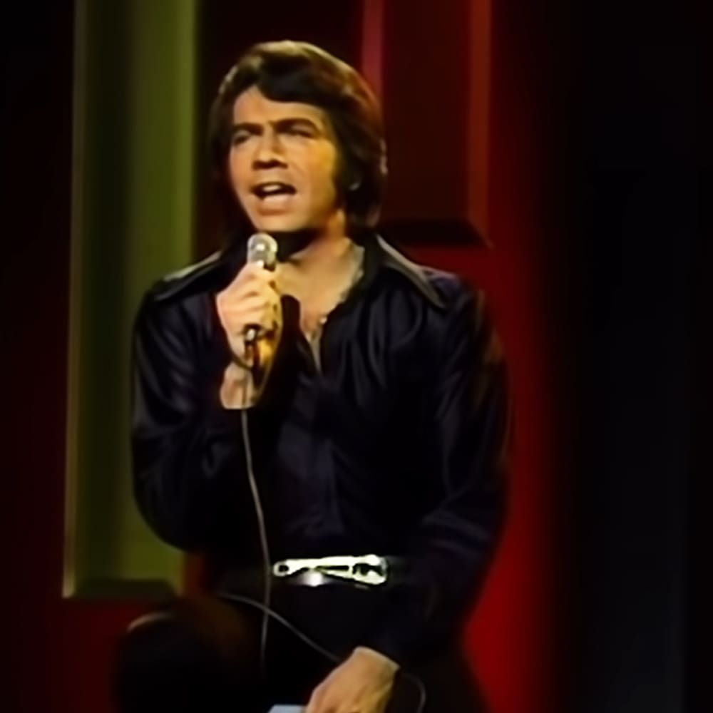 An adult male performer singing into a microphone, wearing a dark-colored leather-like jacket at an indoor event with stage lighting focused on him.
