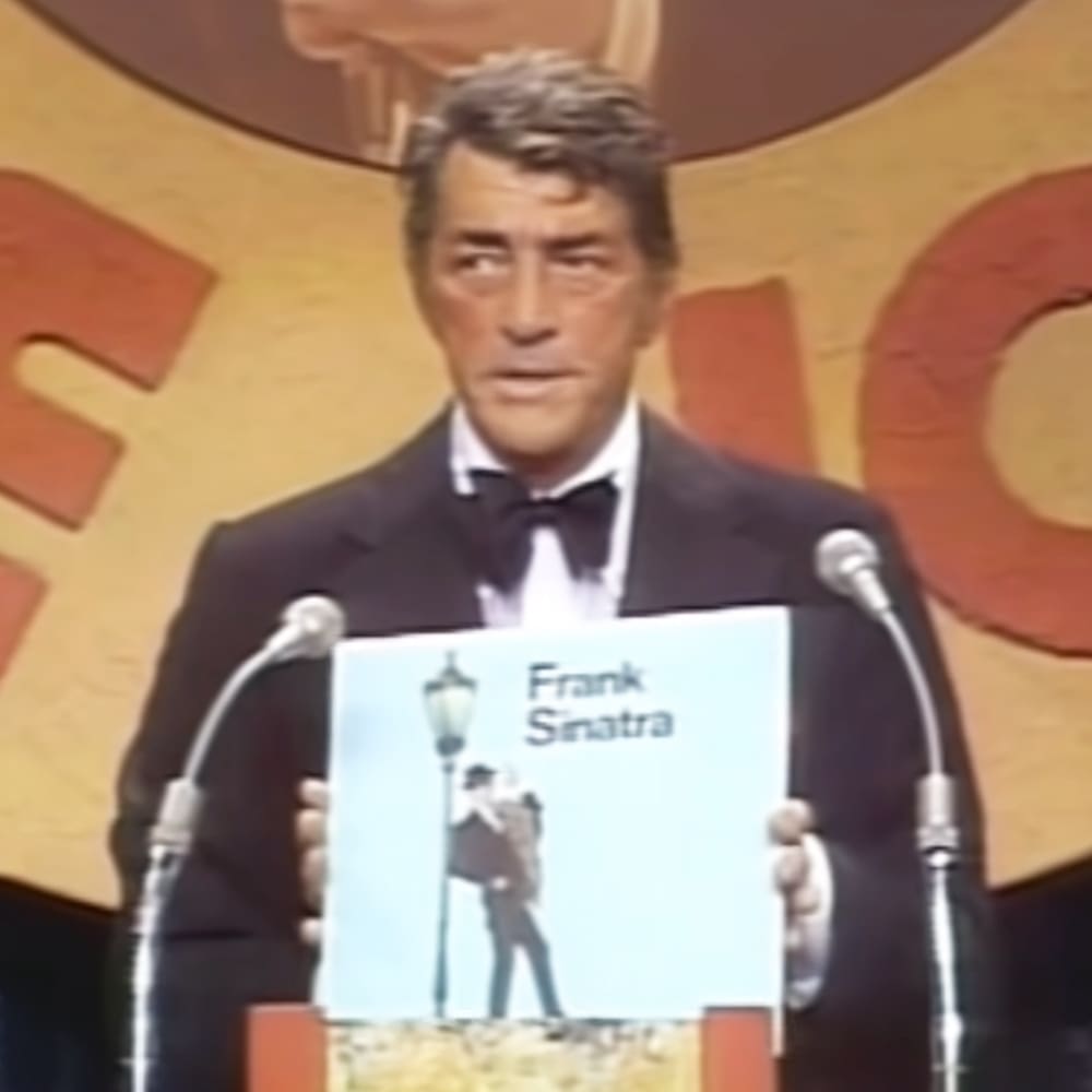 A middle-aged man in a dark suit jacket, white shirt, and black bow tie delivering an acceptance speech at a formal event, possibly an awards ceremony.