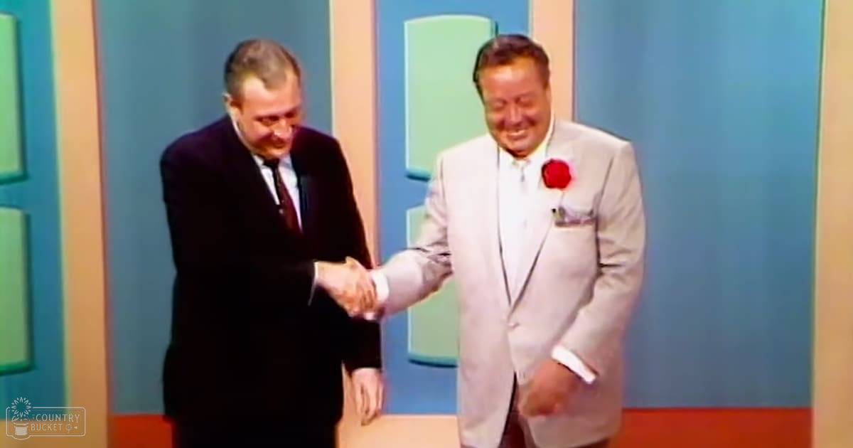 An adult male singer in a dark suit with a subtle patterned tie stands next to another adult male wearing a light gray suit jacket and red boutonniere, set against colorful vertical and horizontal striped panels suggesting a television studio or stage for a formal event.