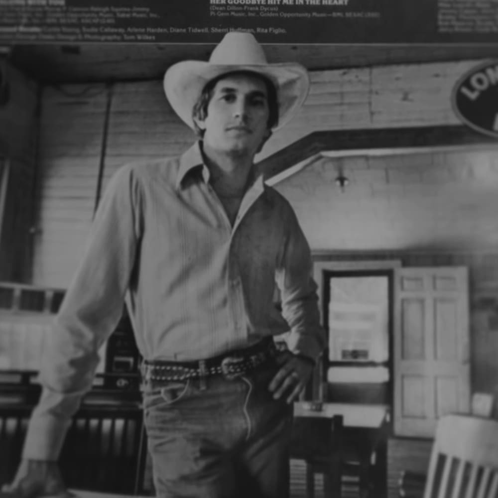 A young adult male wearing a white collared shirt and cowboy hat, seated indoors with wooden furniture and signs in the background.