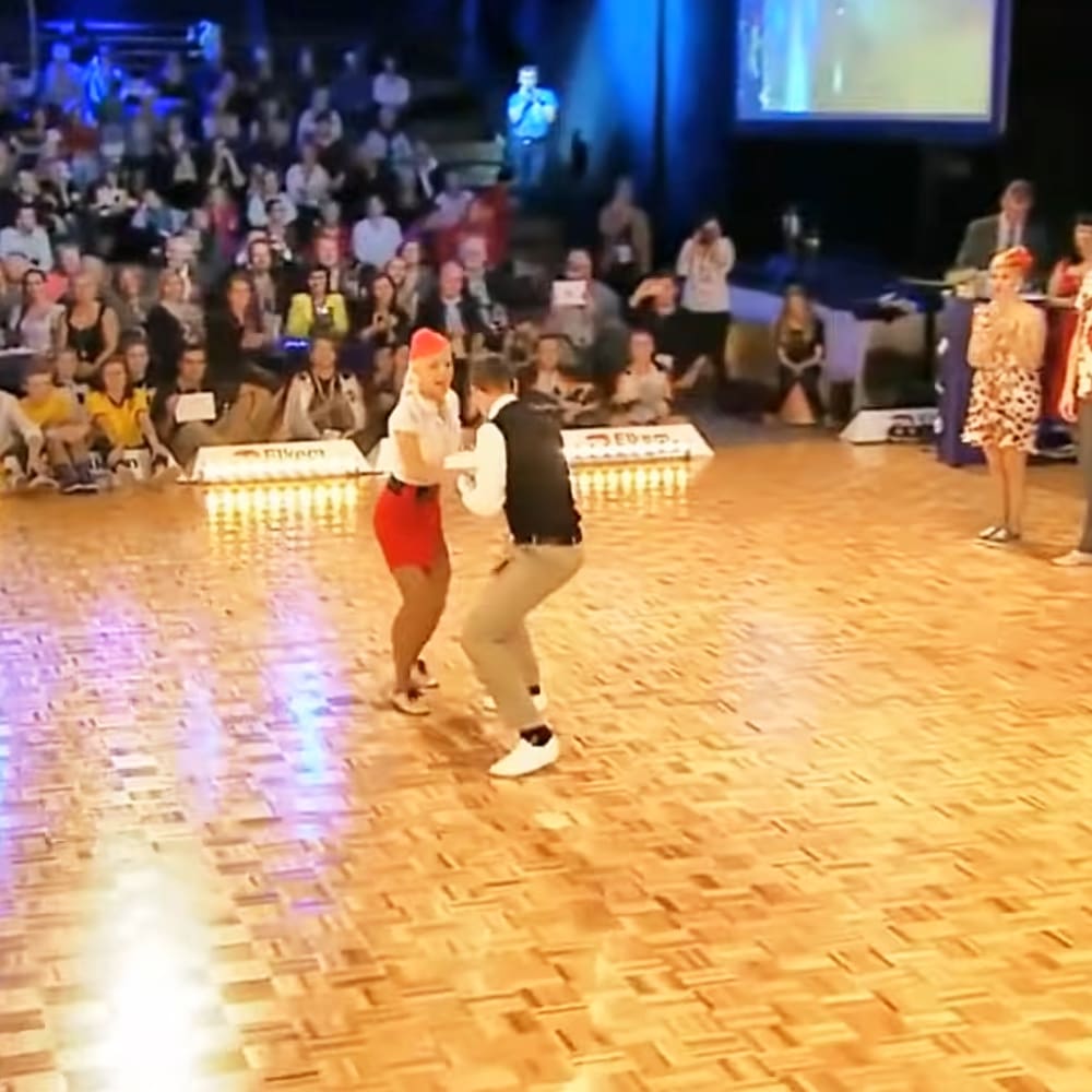 A dance performance in an indoor venue with a young adult female dancer in red shorts and white top performing alongside another young adult male dancer wearing beige pants and dark vest, surrounded by seated spectators in various attire. The setting includes wooden flooring, seating for spectators, and stage lighting.