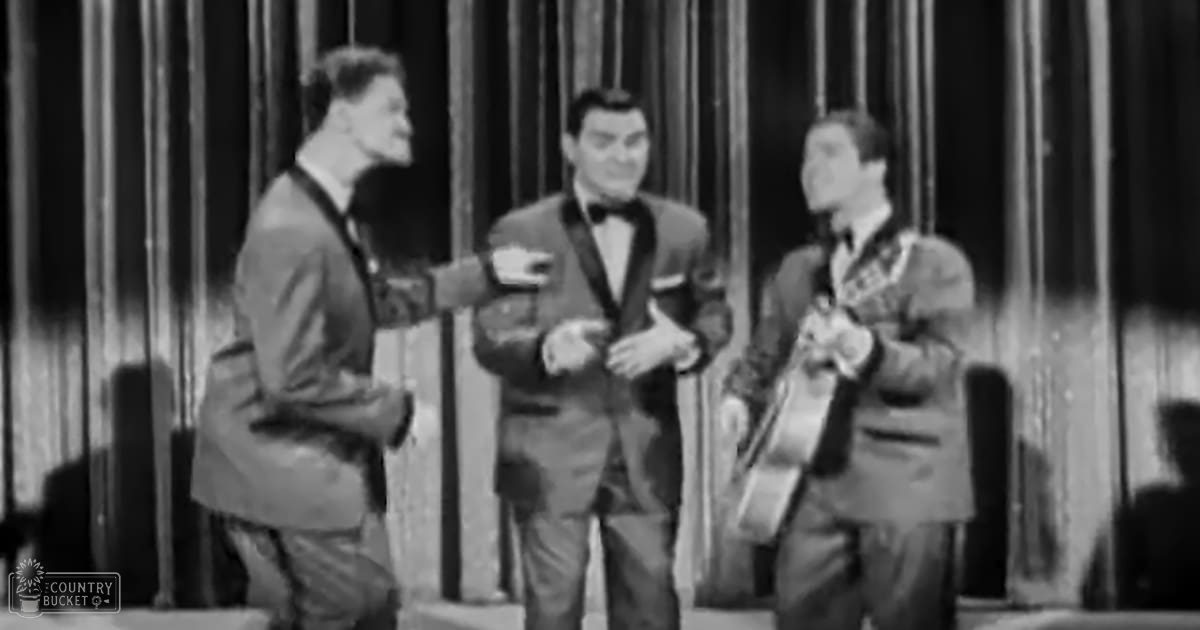 A stage performance scene with three adult male singers dressed formally, each holding a microphone or musical instrument; dark curtains in the background suggest an indoor theater setting.
