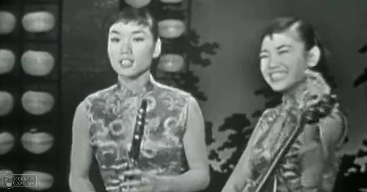 Two young adult females performing a musical act on an ornate indoor stage with traditional-style garments and intricate embroidery, set against a decorative backdrop.