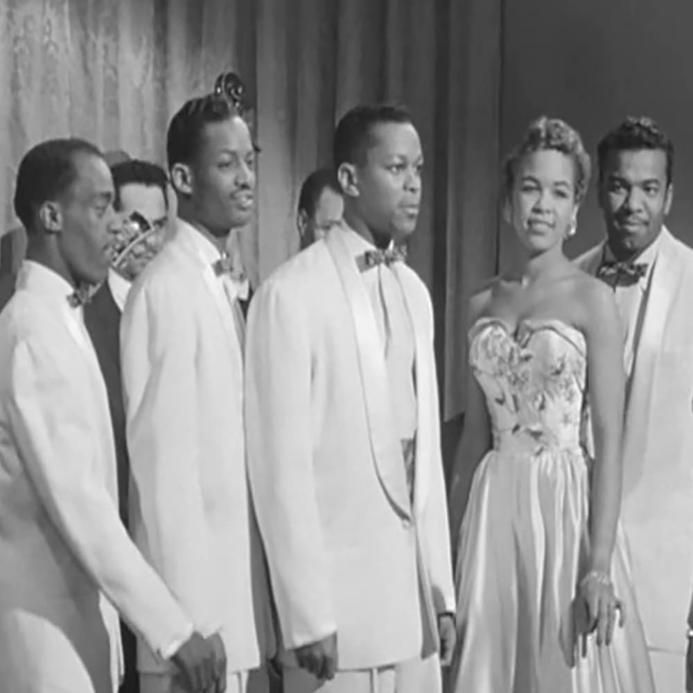 A group of young adult performers in formal attire, possibly from a musical or theatrical production, indoors.