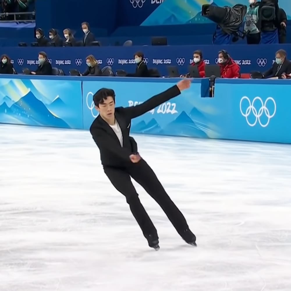 A young adult male figure skater in a black suit jacket, white shirt, and black pants, skating at the Beijing 2022 Winter Olympics indoor ice rink with blue banners and spectators wearing masks.