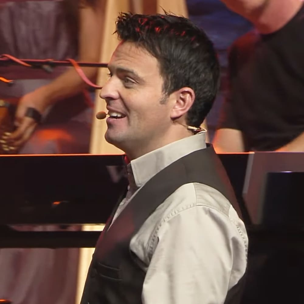 A male adult singer wearing a light-colored shirt with a dark vest over it, performing on stage or in a concert hall setting.