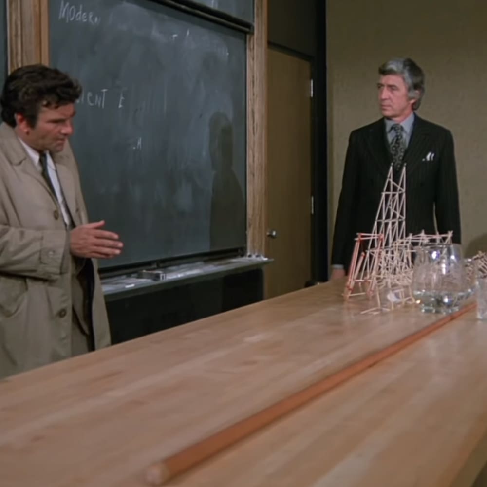 Adult male singer wearing beige trench coat in a classroom setting with older adult male in black pinstripe suit, suggesting an educational environment from the mid-20th century.