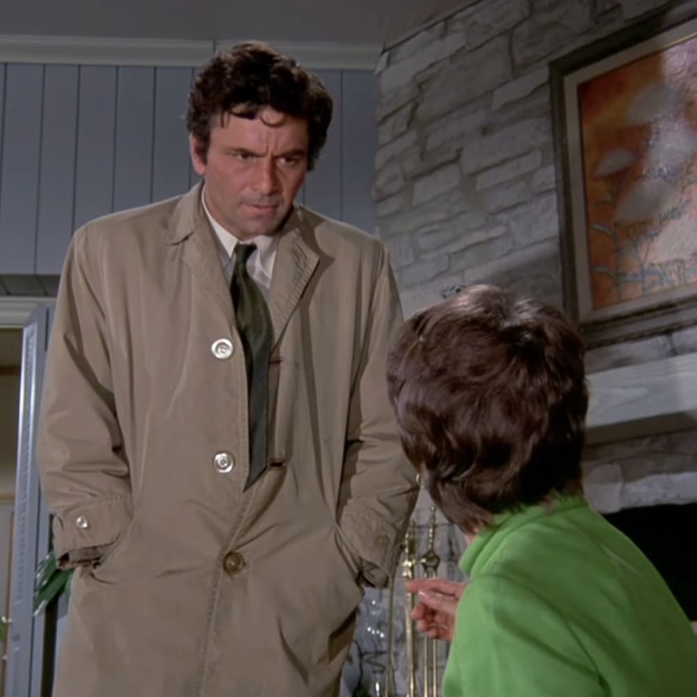 Adult male in beige trench coat with silver buttons, adult female in bright green top indoors near stone fireplace and framed artwork.