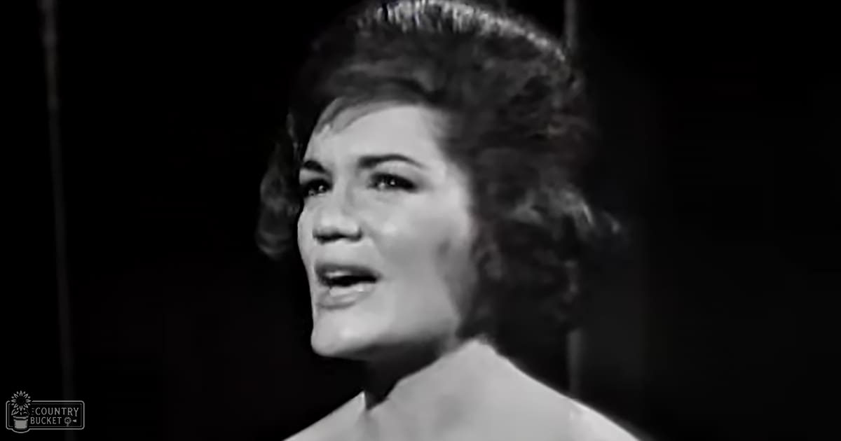 An adult female in an elegant white strapless dress with a scoop neck, likely from the early to mid-20th century, against a dark stage background.