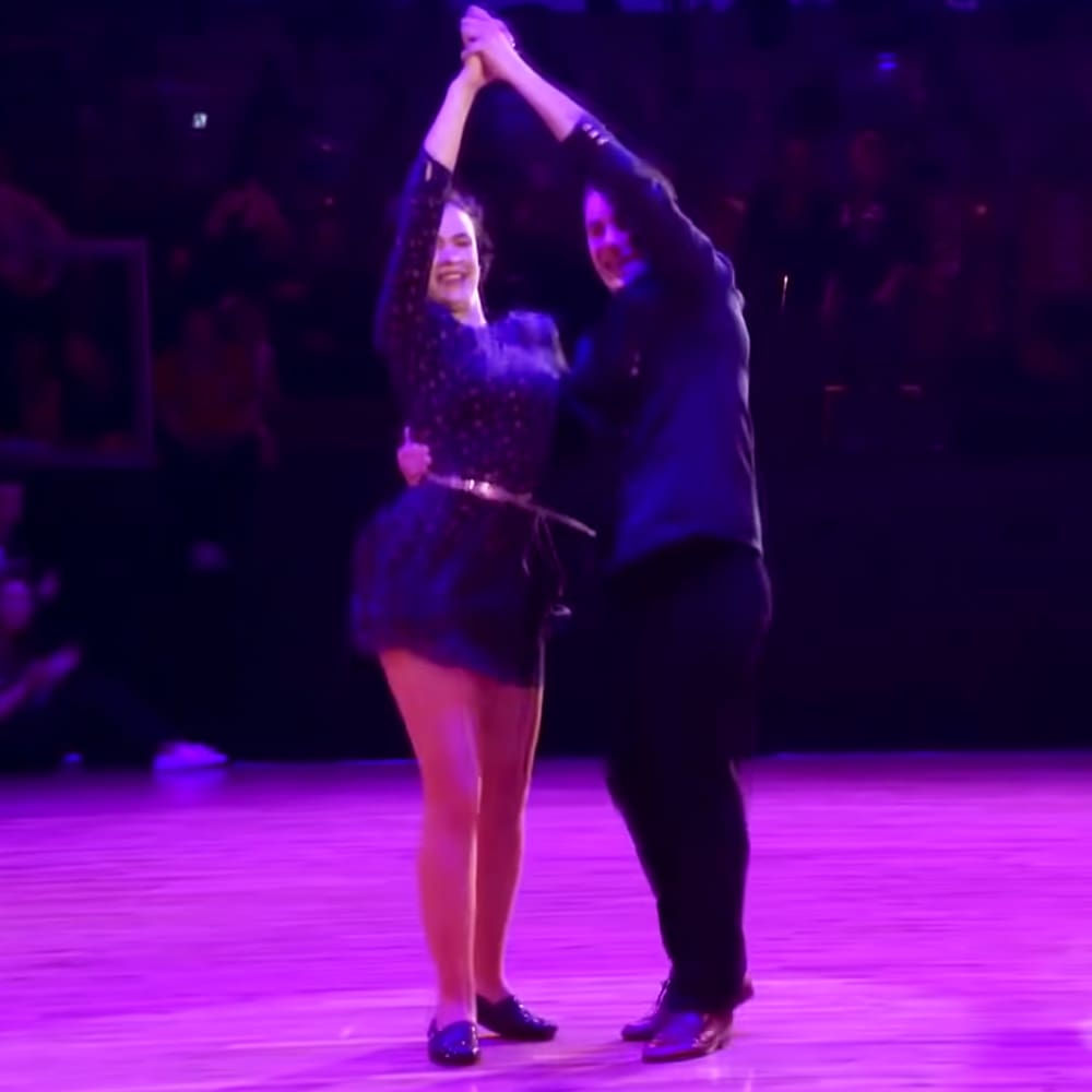 A young adult female dancer in a dark polka-dot dress dances alongside an adult male wearing a dark jacket, at an indoor dance competition with dim purple stage lighting.