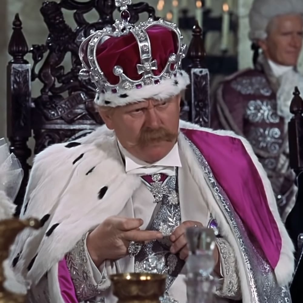 A man dressed as a king in historical attire, seated on an ornate wooden throne surrounded by formally dressed individuals in what appears to be a castle or royal hall.