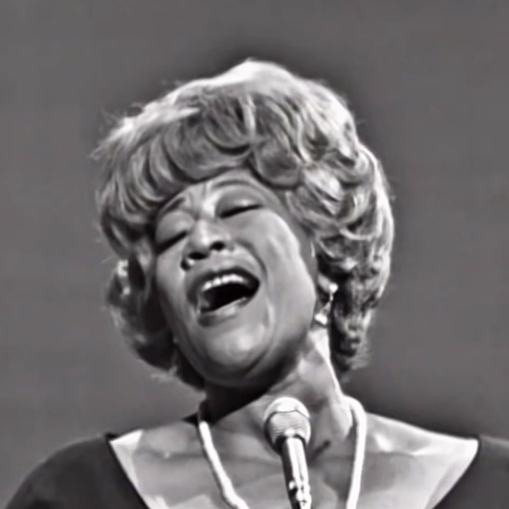 A middle-aged woman singing into a microphone on an indoor stage with minimal background detail.