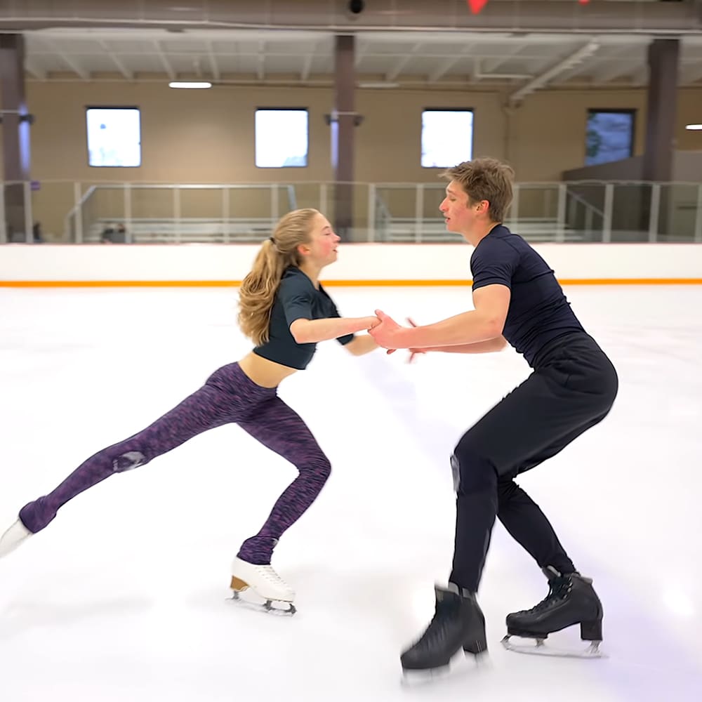 A young adult female in purple leggings skating with a young adult male in an indoor ice rink.