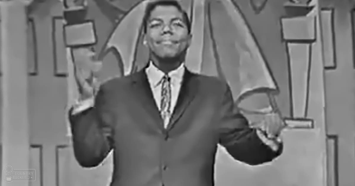 An adult male performer wearing a dark suit jacket with a patterned tie stands on stage against a geometric backdrop, likely from a mid-20th century television show or variety program.