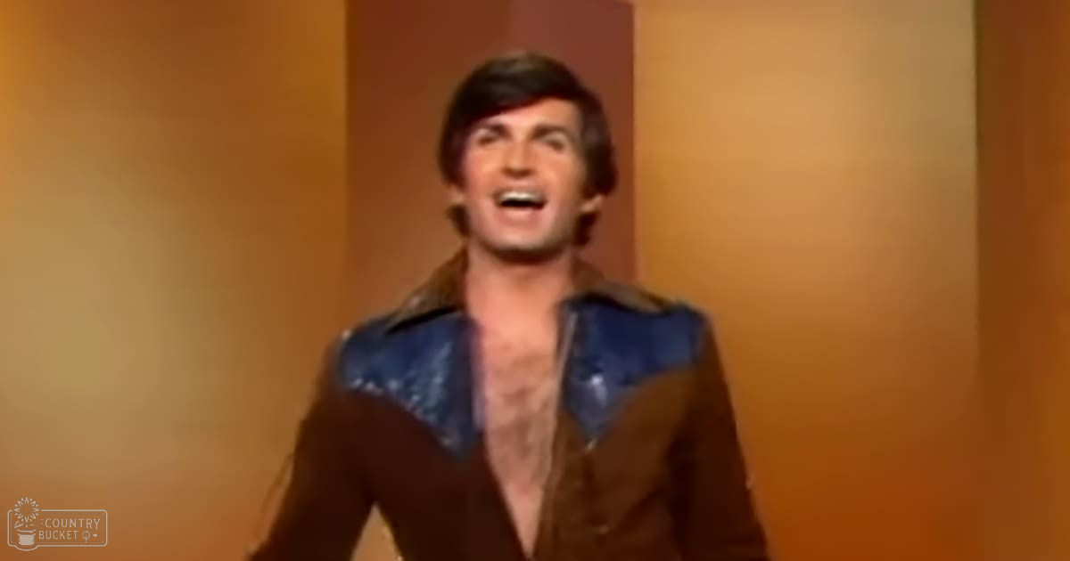 A young adult male wearing a brown jacket with blue accents on the shoulders in an indoor setting with warm lighting.